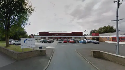 Google The Geoff Moulder Leisure Complex - a single-storey brick and metal building with a red roof. A car park with several cars is in front, along with a pedestrian crossing. A white sign with blue text marks the entrance. Trees and a cloudy sky are visible in the background.