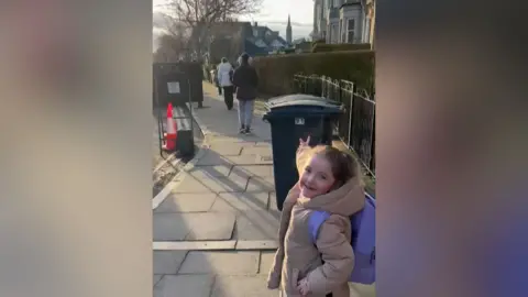 A small girl on a pavement in the street in a beige jacket pointing at the sky.