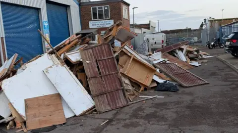 Tony Milson A large pile of wooden waste dumped on an industrial estate outside a garage which has its blue shutters down. There is a van and a motorbike in the background and the back of a car on the right of the picture.