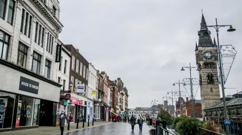 Darlington High Street has a large clocktower on the right of a row of shops. Several shoppers are walking up the paved street. A row of street lights can be seen leading down the street on the right.