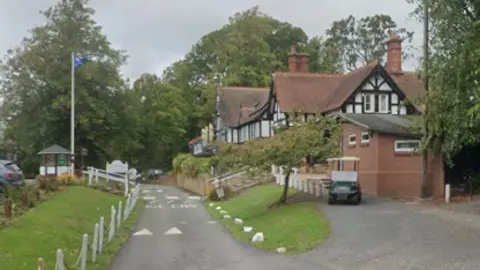 Google A half-timbered building with red-tile roof overlooks a road with white chain-and-post fencing along the grass verge, and a blue flag on a pole opposite. 
