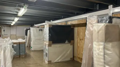 Supplied The inside of a warehouse or storage facility, where mattresses are stacked up in rows.