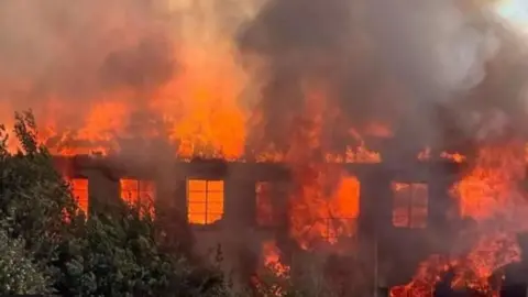 The Saxonvale fire - there are big orange flames and lots of thick, grey smoke. Through the flames, the remnants of the derelict building can be seen.