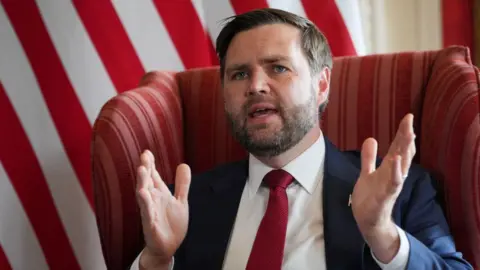 Reuters JD Vance sits in a chair, wearing a suit and tie. He has his mouth open and is speaking, with his hands in the arms to emphasise a point.