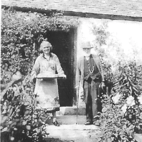 Galbraith A black and white picture of a woman with a tea tray standing at a cottage door and a man with a walking stick in a hat and trousers and waistcoat