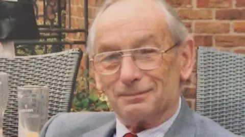 Simon Smith An elderly man with thinning grey hair and glasses smiles at the camera. He sits in front of a red brick wall with a glass visible to his right and the backs of rattan chairs.