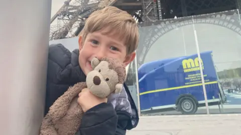 Family handout A young boy peeking round the corner of a grey post smiling in a cheeky way and holding a brown monkey teddy up to his cheek. He has dark blond hair and a fringe and is wearing a black coat. The bottom of the Eiffel Tower is in the background, as is a reflection of a blue van.