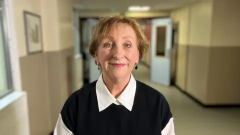 Prof Dwyer stands in an empty hospital corridor- she is wearing a white shirt and a black sleeveless jumper. She is smiling and looking directly at the camera. 