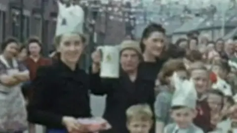 Yorkshire & North East Film Archive Vera McLoughlin standing next to a table laid out for the street party. Children are sat at the table before her. She is wearing a hat and a black coat. She is holding a white jug in the air to cheer.