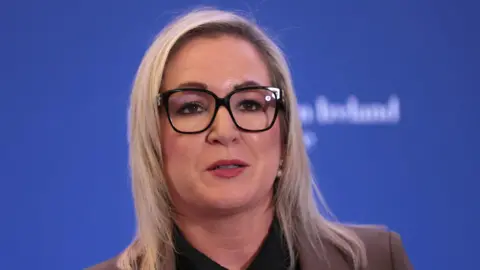 PA Media Michelle O'Neill. She has shoulder length blonde hair, wearing glasses and a brown blazer with brown blouse. Behind her is a blue background.