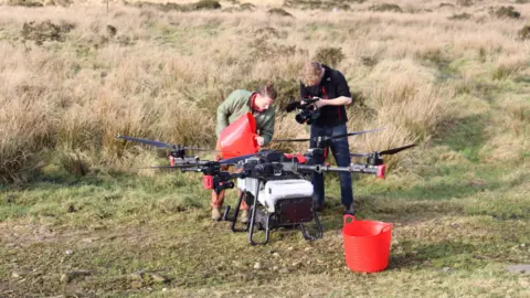 Here Now Films Two people standing on moorland load seeds into a large drone. One person is holding a large red plastic bucket and is pouring seeds into the drone. The other person is filming them on a video camera.