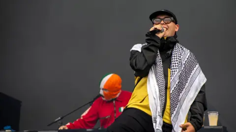 PA Media Liam Og O Hannaidh (Mo Chara) from Kneecap performs onstage - he is wearing a keffiyeh, glasses and a baseball cap, while bandmate DJ Provai is behind him on the decks.