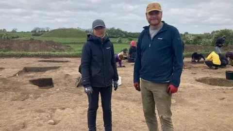 A man and a woman standing at an archaeology dig site. Both are wearing blue jackets, gloves and caps. There are a number of excavations in the background with people looking into them.