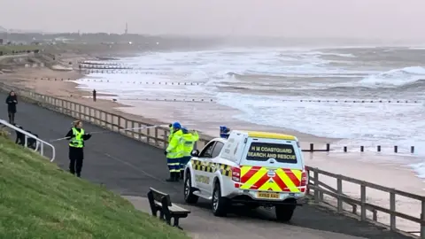 BBC Coastguard van at beach with grey skies and rough seas behind a line of police tape.