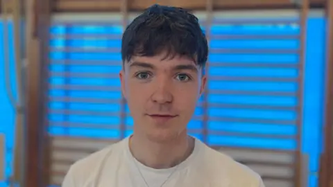 Sean is looking at the camera. He has short brown hair and is wearing a plain white t-shirt.