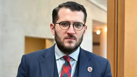 Getty Images Paul Sweeney, a man in a dark blue suit with a chequed multi coloured tie. He has dark hair, a beard and glasses.
