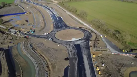 Essex Highways An aerial shot from above showing a newly built road with a roundabout in the centre of the picture. To the right of frame is a green field, to the left a smaller road leads off to muddy ground.