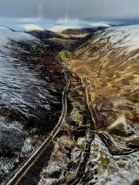 Suzi Watson An aerial view of a road cutting through a valley between two snowy hillsides.