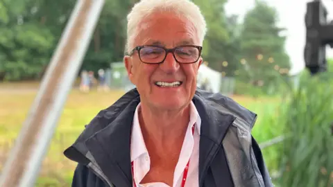 Jon Wright/BBC A man with white hair stands on a festival stage with reeds and trees behind him.