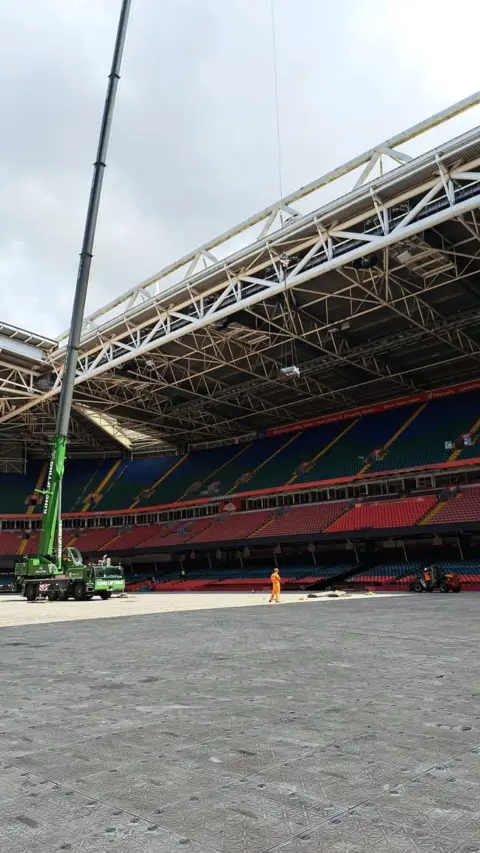 WRU A huge green crane can be seen lifting something which is in mid air. The crane is set on a hard flooring in the bowl of a sport stadium.