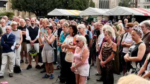 Mousumi Bakshi/BBC A crowd of people standing in a market square looking on as they are addressed by a speaker at a vigil.