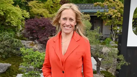 Getty Images Sophie Raworth with long blonde hair and an orange coat with orange jumper underneath stands in front of a garden scene