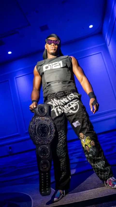 Jordan Lees Pro wrestler DeReiss Gordon, a muscular man poses confidently in a room bathed in blue lighting. He wears sunglasses displaying "DRAGO," a black stab proof vest with "0121" printed on it, a black durag cap, and black cargo pants featuring graffiti-style writing. He holds a large wrestling belt. 