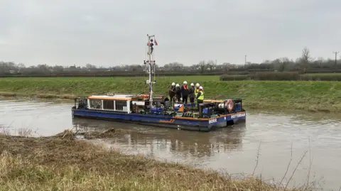 A blue boat with orange piping is in the middle of the shot on the river. A group of people in hi-vis jackets, buoyancy aids and hard hats are on the boat. Behind it is a green field.