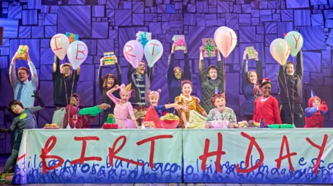 8 child actors are on stage in different fancy dress costumes, they're celebrating a birthday with balloons, food and presents. They are all stood in a different pose during the scene with the actors playing their parents stood behind them holding presents in the air. 