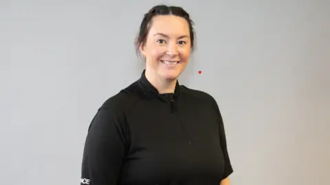 Staffordshire Police A woman with tied-back dark hair and a black police shirt smiles as she stands in front of a light grey wall.