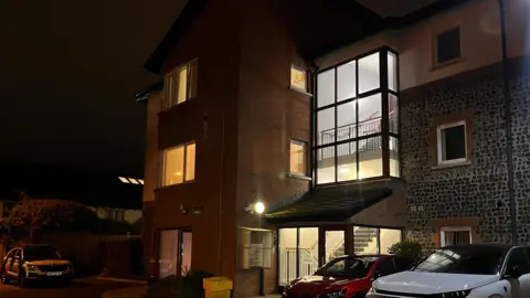 A police car and red and white cars are parked outside a red brick block of flats which is lit up