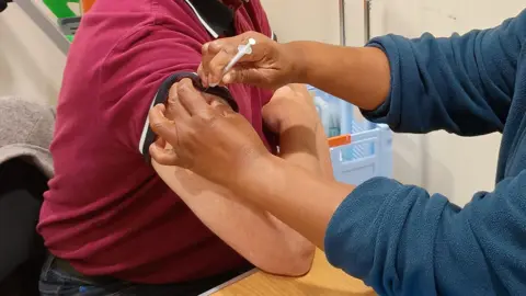 Cambridgeshire & Peterborough Integrated Care System A man is sat on the left with his arm on the table. He is wearing a red polo shirt and someone is putting a vaccine in his upper arm. A person is on the right who is giving the vaccine, and is wearing a blue jumper.