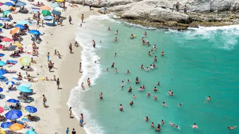 An aerial view of beachgoers swimming in the Tyrrhenian Sea or sheltering under umbrellas on the beach in Tropea, Italy.