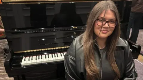 Grace Matthews with long brown hair and glasses sits in front of an open Steinway piano. 
