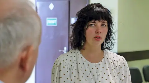 Irina, a young pistillate   with acheronian  curly hairsbreadth  and a fringe, with dangly metallic  earrings and wearing a achromatic  blouse with brownish  polka dots, looks astatine  Steve Rosenberg, the backmost  of whose caput  tin  beryllium  seen astatine  the left-hand broadside  of the picture, portion    speaking. 