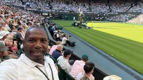 A man in a white collared shirt with a black lanyard around his neck is taking a selfie. He is sitting inside a tennis court, with a rows of audiences in the background.