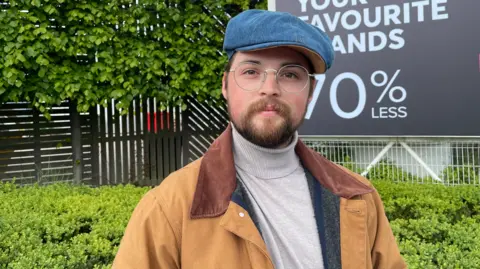 Elizabeth Baines / BBC A man with a blue cap wears a brown jacket. He has silver spectacles and stands infront of a green hedge and a billboard.