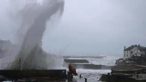 BBC Weather Watchers/MaxieHJ This picture shows a plume of sea spray shooting high into the air, towering above the buildings and vehicles nearby. A harbour is churning with rough, fast‑moving water, and the sky looks grey and unsettled. A few parked cars and a piece of machinery sit on the quayside.