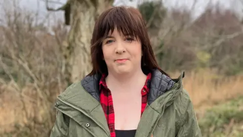 BBC Michelle Martin wearing a green coat and plaid shirt looking at the camera with a slight smile. She is standing outside with some trees in the background.