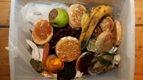 Food waste is photographed from above in a bin. The bin is stood on floorboards within a white bin liner.