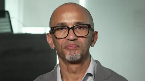 Desmond, now in his fifties, looks directly into the camera. He is wearing glasses and a light grey suit jacket and shirt. 