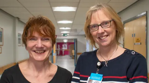 Two women stand side by side in a hospital corridor. Mel Milner has a pixie cut with copper-ginger hair and wears a black T-shirt. Sarah Redmond has chin-length blonde hair, wears a navy sweater with red and yellow stripes, and black glasses. Both women are smiling at the camera.