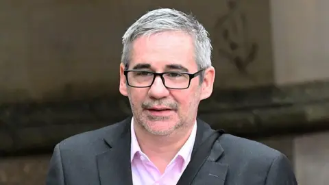 Iain Robertson wears a grey suit and pink shirt. he has short grey hair and wears dark glasses. He is walking outside a building