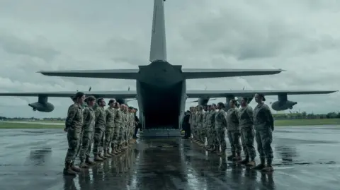 Military personnel form two lines, facing each other, to the rear of a military aircraft which has its ramp down at the bottom. It is a grey and wet day. The servicemen and women have their hands placed behind their backs. 