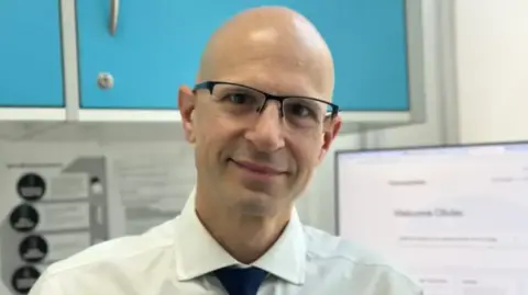 Olivier Picard Olivier Picard smiles at the camera while standing in a pharmacy. His head is shaven and he wears glasses, a white shirt and a dark tie.