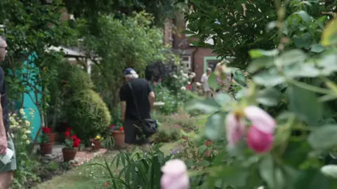 A wide shot of a green garden, behind is a man wearing all black and a cap