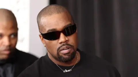 Headshot of West who wears a silver chain and sunglasses. There is a man behind him who is blurred 
