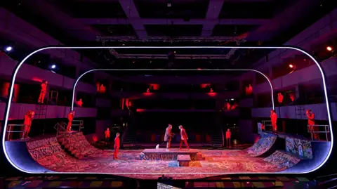 A theatrical production, showing a box like structure in a theatre, with several actors standing on it. It represents a skatepark. There is painted graffiti on the ground. Rows of seats can be seen around the box.