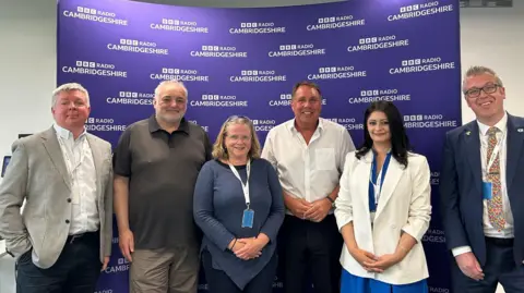 Andrew, a man with grey hair -wearing a white shirt and a blazer, standing next to Chris Hogg, a man in a brown T-shirt, Heather, a woman - wearing a blue jumper, Chris Mann, wearing a white shirt, Shabina, a woman - wearing a white jacket and a blue dress, John, a man - wearing a blue suit. They are standing in front of a purple BBC Radio Cambs board, smiling for the camera. 