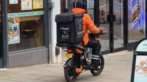 Man in orange jacket rides electric bike through city centre with large black food delivery rucksack on his back
